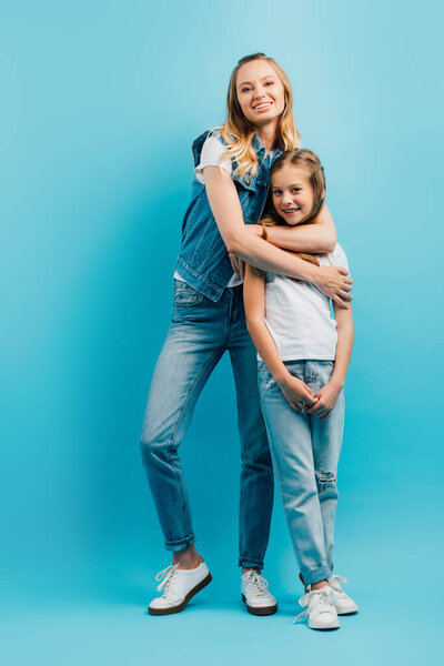 молодая женщина в джинсовой одежде обнимает дочь в белой футболке и джинсах, глядя на камеру на синем