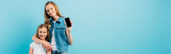 ブルーに隔離された娘を受け入れながら 空白の画面を持つスマートフォンを示すデニムベストの母親の水平画像 — ストック写真