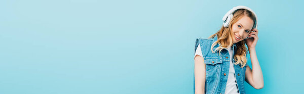 website header of woman in denim clothes touching wireless headphone while looking at camera isolated on blue