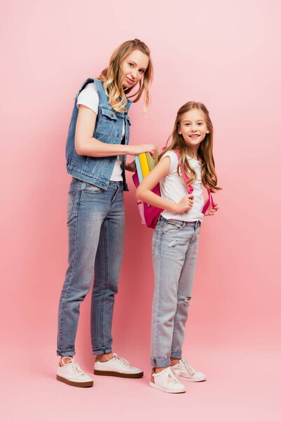 Вид в полный рост молодой женщины, надевающей рюкзак на спину дочерям, глядя в камеру на розовом фоне