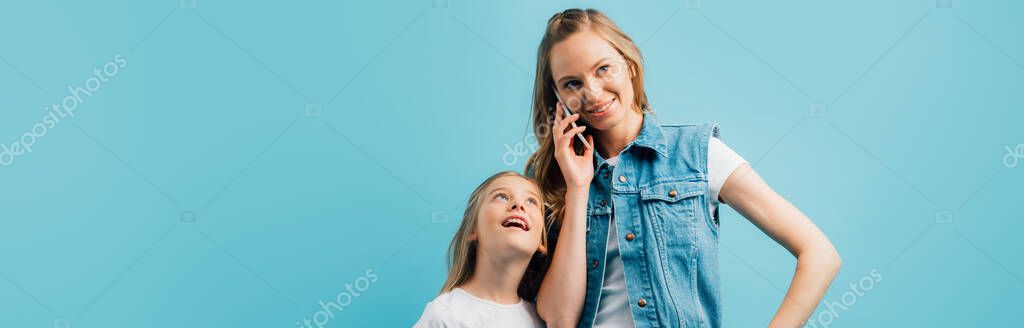 Panoramic shot of young woman in denim vest talking on smartphone near curious daughter isolated on blue — Stock Image Panoramic Shot Young Woman Denim Vest Talking Smartphone Curious Daughter Royalty Free Stock Images