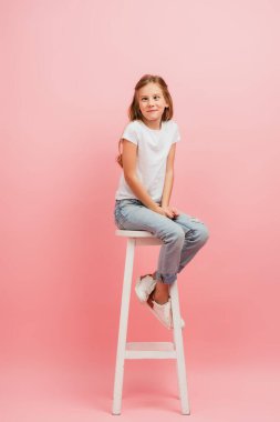 kid in white t-shirt and jeans grimacing and squinting while sitting on high stool on pink clipart