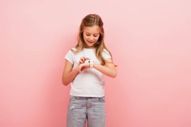 Beyaz tişörtlü ve kot pantolonlu çocuk pembe üzerinde izole edilmiş akıllı seke dokunuyor.