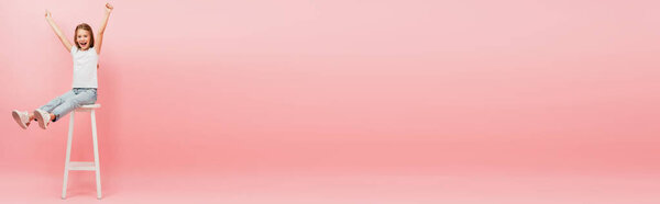 девушка в белой футболке и синих джинсах сидит на высоком стуле с протянутыми ногами и показывает жест победителя на розовый, панорамный снимок