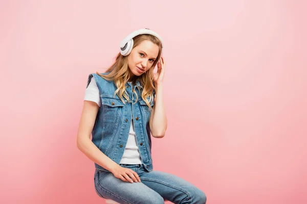 Młoda Kobieta Dżinsowych Ubraniach Dotykająca Bezprzewodowych Słuchawek Siedząc Odizolowana Różowo — Zdjęcie stockowe