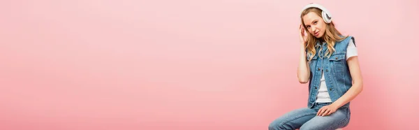 ピンクに隔離されたままワイヤレスヘッドフォンに触れるデニム服の若い女性のパノラマの概念 — ストック写真