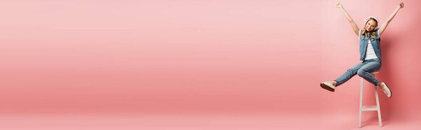 горизонтальное изображение молодой женщины в беспроводных наушниках, сидящей на высоком стуле и показывающей жест победителя на розовом