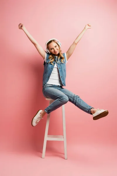 興奮した女性ですワイヤレスヘッドフォンピンクの上に高いスツールに座っている間に勝者のジェスチャーを示す — ストック写真