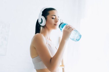 Kulaklıklı kadın evde spor şişesinden su içiyor.  