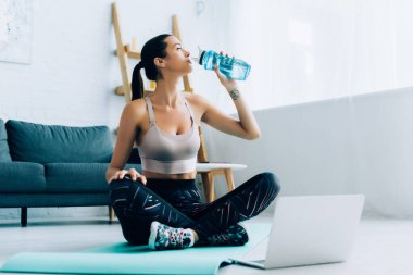 Dizüstü bilgisayarın yanında fitness minderinde otururken kadın sporcunun içme suyu seçici odağı 