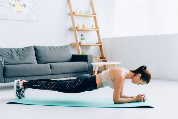 Side view of sportswoman standing in plank on fitness mat in living room 