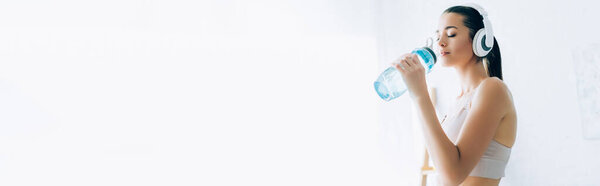 Panoramic shot of sportswoman in headphones drinking water at home 