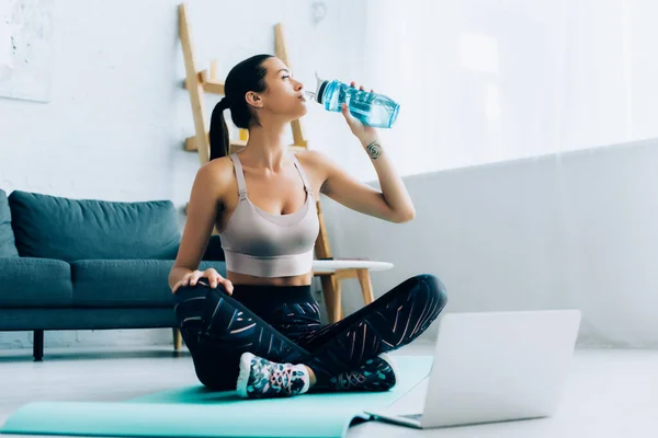 女运动员坐在笔记本电脑旁边的健身垫上喝水时的选择性焦点 — 图库照片
