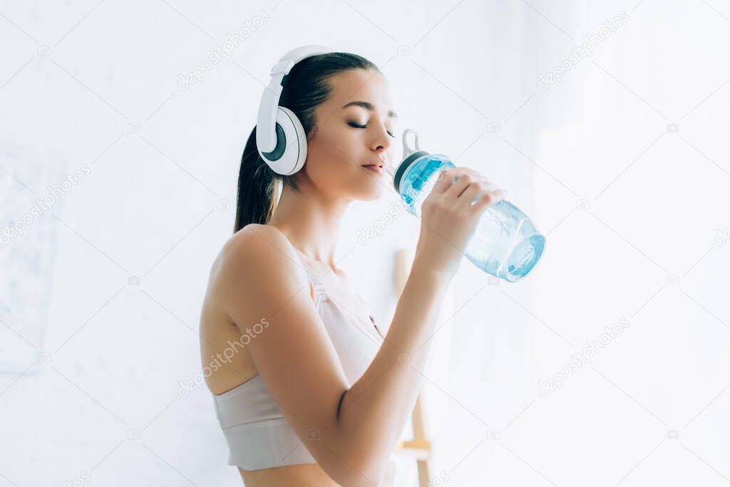 Sportswoman in headphones drinking water from sports bottle at home  