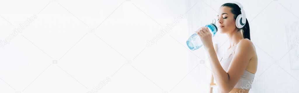 Panoramic shot of sportswoman in headphones drinking water at home 