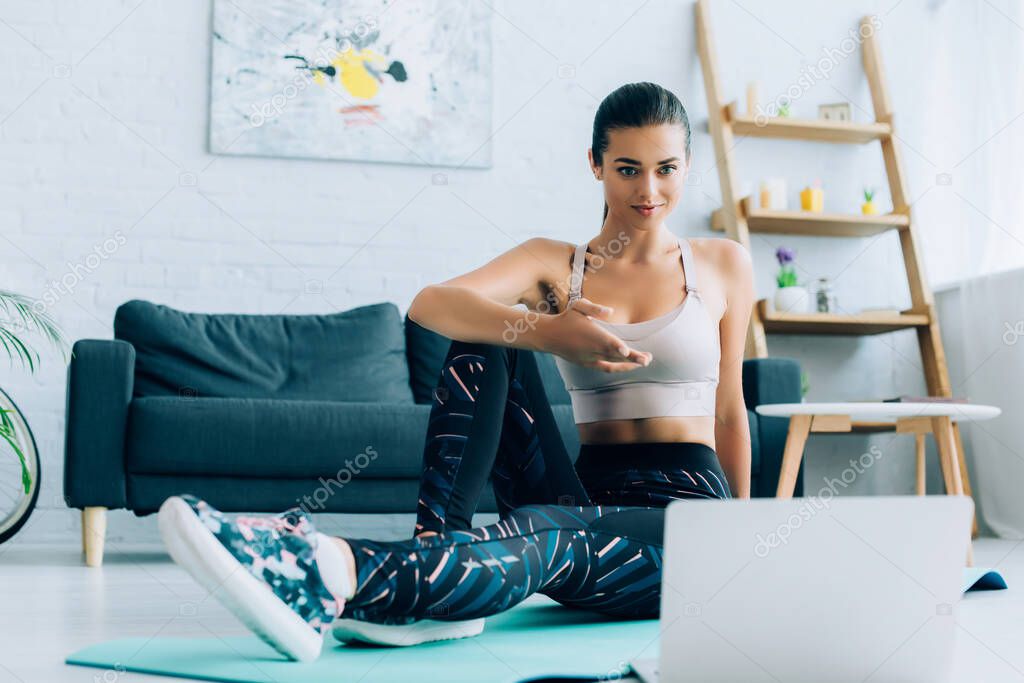 Selective focus of sportswoman pointing with hand at laptop while sitting on fitness mat at home 
