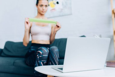 Laptopun seçici odak noktası sehpa ve kanepede direnç bandı tutan sporcu kadın. 