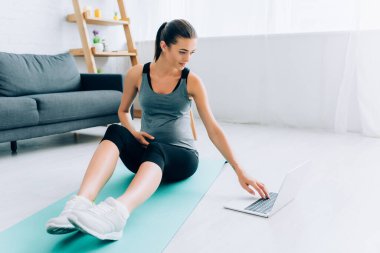 Hamile bir sporcunun evdeki spor minderinde dizüstü bilgisayar kullanmasının seçici odağı 