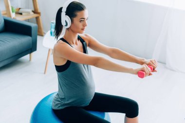 Kulaklıkla müzik dinlerken dambıl ve fitness topu taşıyan hamile bir sporcu. 