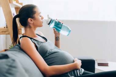 Hamile bir sporcunun karnına dokunup spor şişesini kanepeye koyması.  