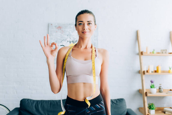 Brunette sportswoman with measuring tape around neck showing okay gesture at home 