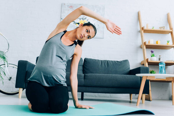 Pregnant sportswoman stretching arm while sitting on fitness mat at home 