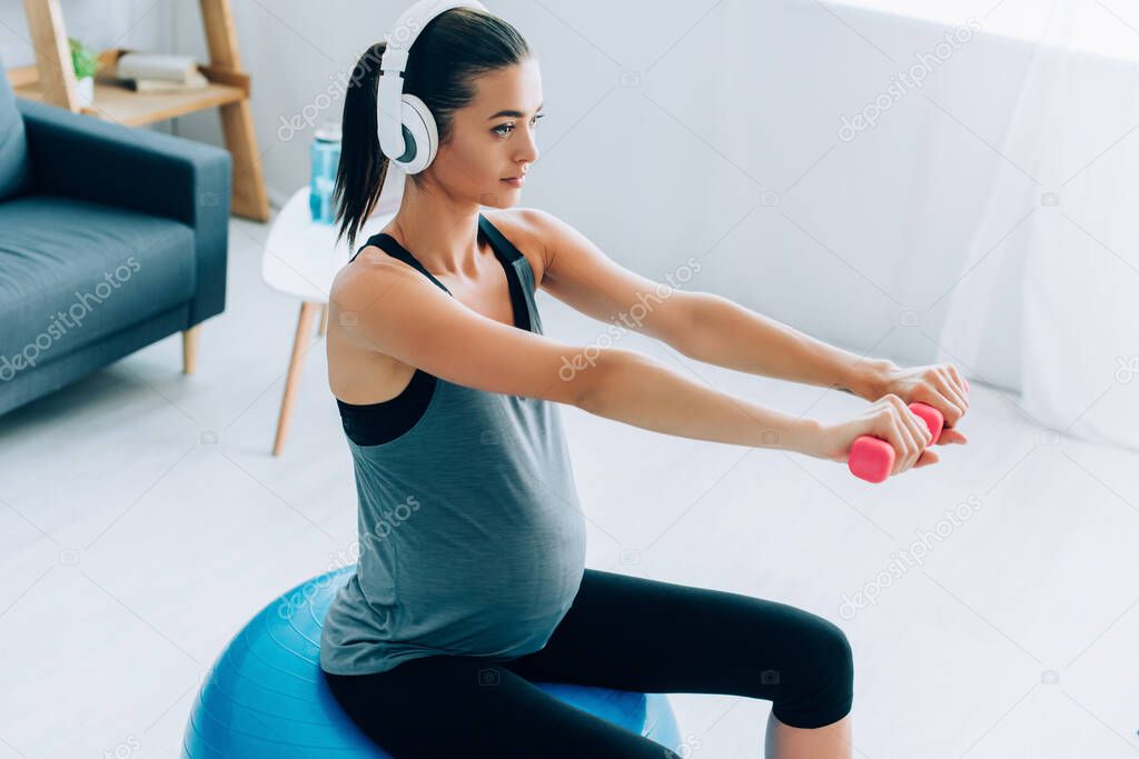 Pregnant sportswoman training with dumbbells and fitness ball while listening music in headphones 