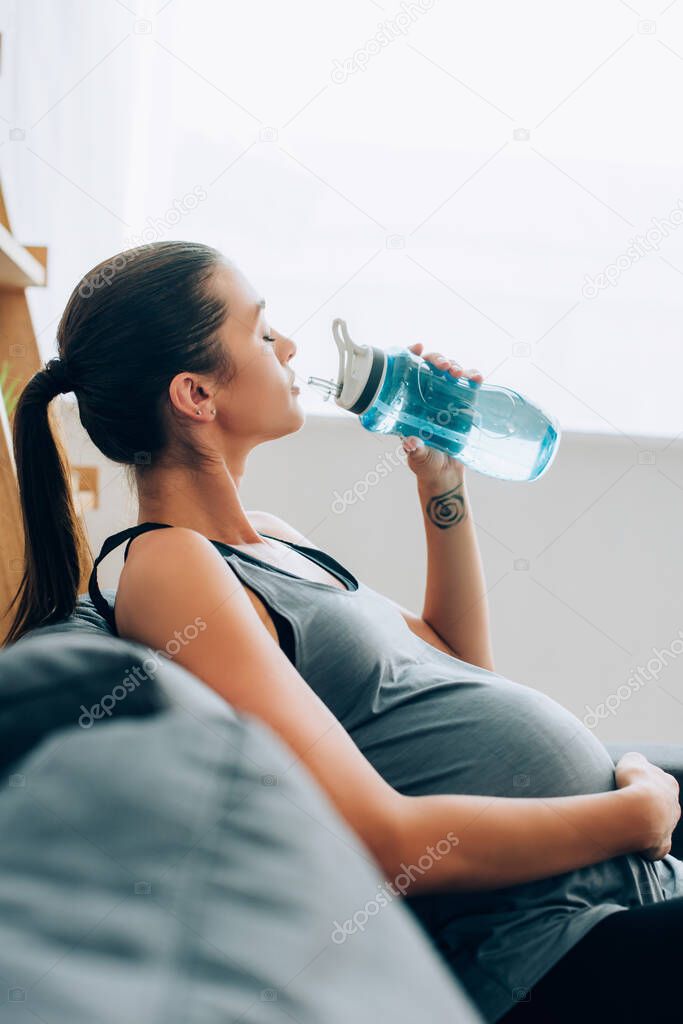 Selective focus of pregnant sportswoman drinking water while sitting on sofa at home 
