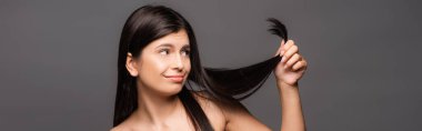 naked skeptical brunette long haired woman looking at split ends isolated on black, panoramic shot clipart