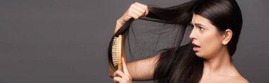 naked shocked brunette woman brushing hair isolated on black, panoramic shot clipart