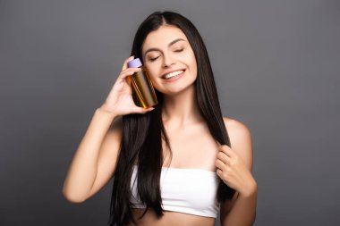 brunette woman holding hair oil in bottle and smiling isolated on black clipart