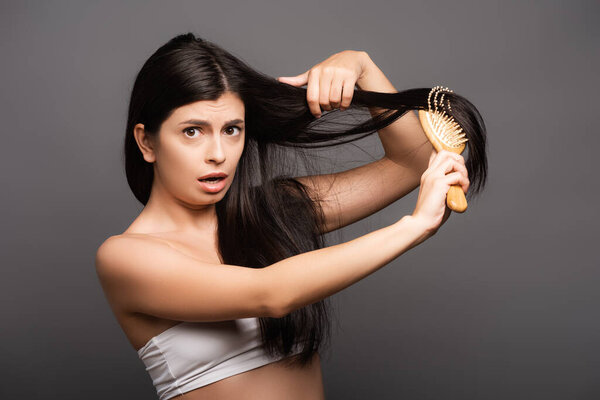 шокированная брюнетка женщина расчесывая волосы изолированы на черный