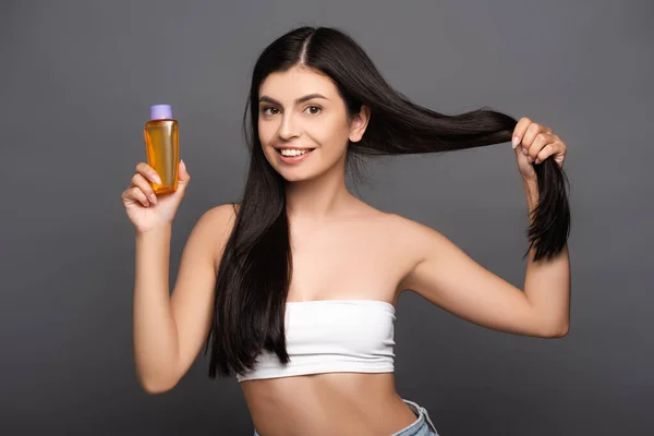 brunette woman holding hair and oil in bottle isolated on black