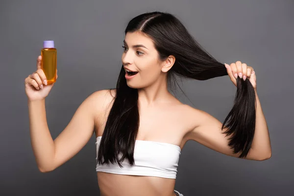 shocked brunette woman holding hair oil in bottle isolated on black