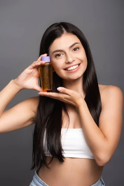 brunette woman holding hair oil in bottle and smiling isolated on black