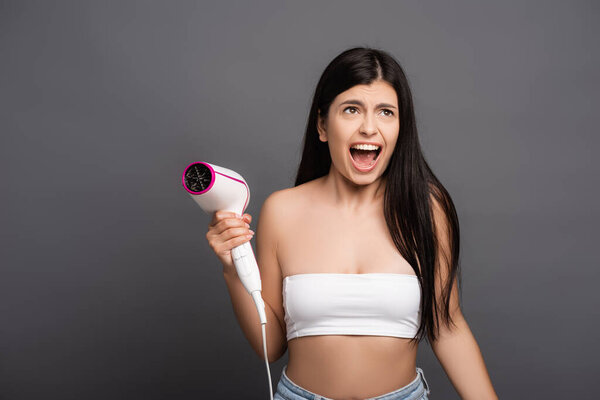 brunette woman holding hair dryer and yelling isolated on black