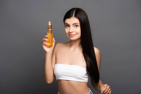 brunette woman holding hair oil in spray bottle and smiling isolated on black