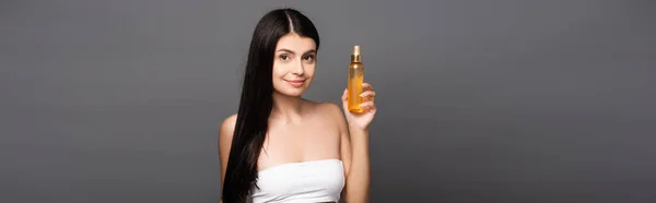 brunette woman holding hair oil in spray bottle and smiling isolated on black, panoramic shot