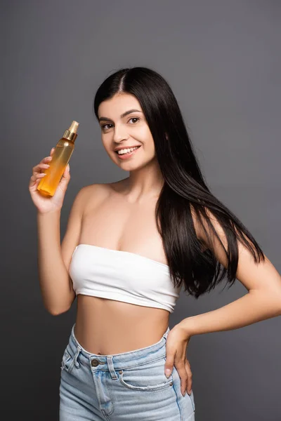 brunette woman holding hair oil in spray bottle and smiling isolated on black