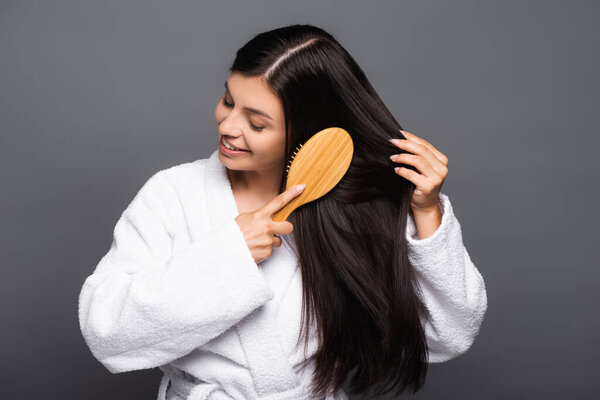 brunette woman in bathrobe brushing hair and smiling isolated on black