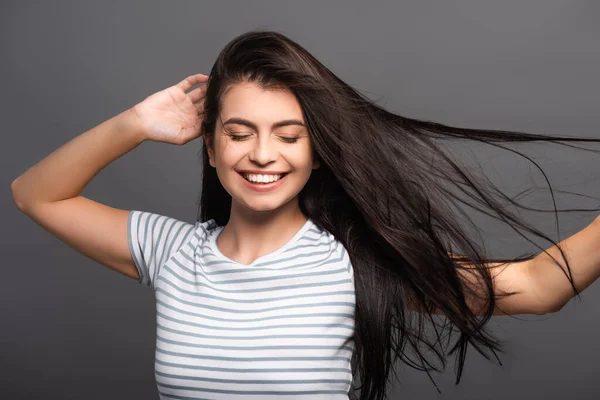 brunette woman with closed eyes smiling and touching hair isolated on black