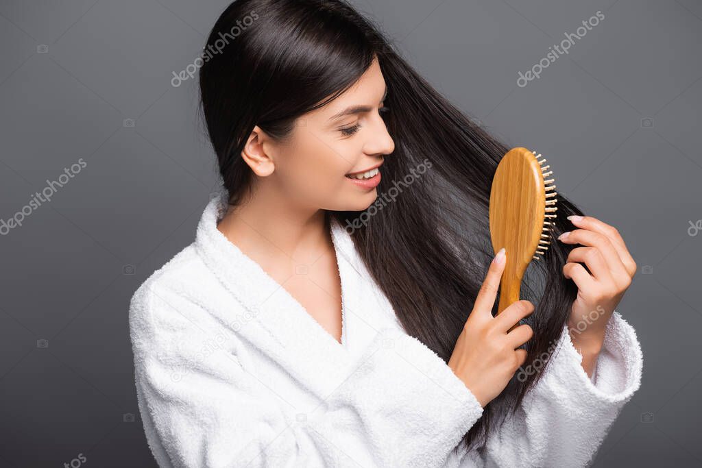 brunette woman in bathrobe brushing hair and smiling isolated on black