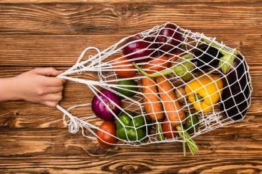 cropped view of woman holding string bag with fresh ripe vegetables on wooden table clipart