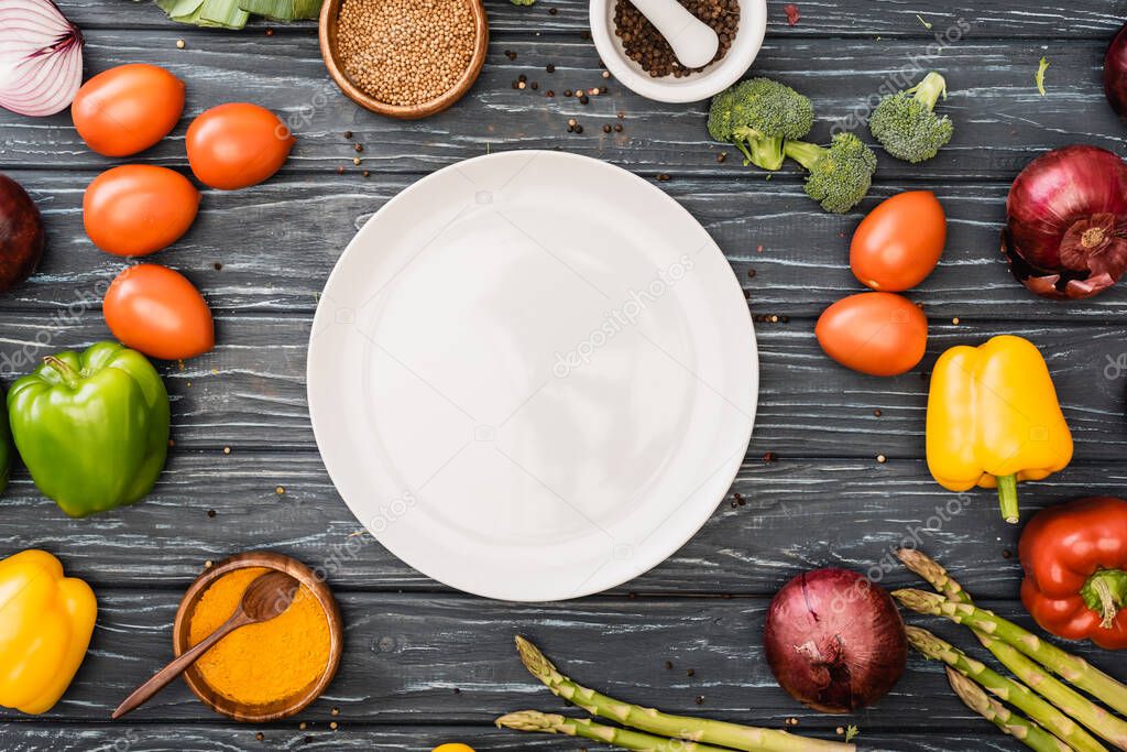 top view of fresh colorful vegetables and spices near empty plate on wooden surface