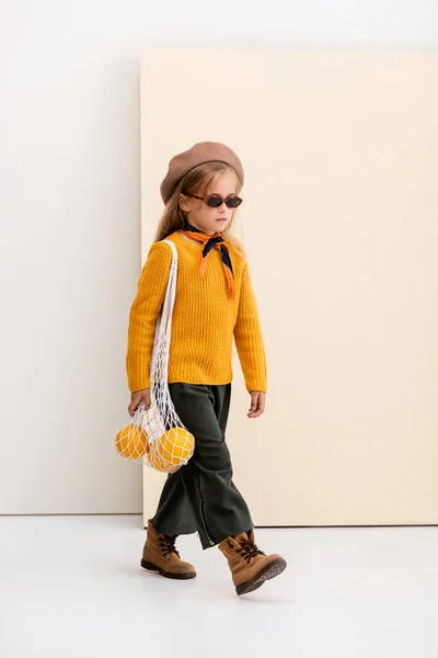 秋の衣装を着たファッショナブルなブロンドの女の子とサングラス歩くととともにグレープフルーツで文字列バッグでベージュと白の背景 — ストック写真