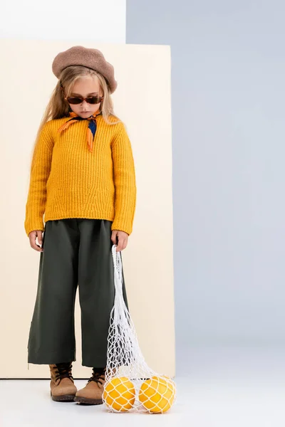 ファッショナブルなブロンドの女の子で秋の衣装とサングラスポーズでグレープフルーツで文字列バッグ上のベージュと白の背景 — ストック写真