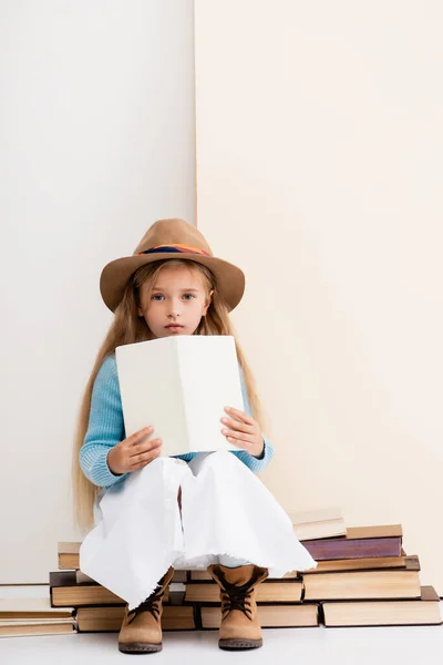 ファッショナブルなブロンド女の子で茶色の帽子とブーツ 白いスカートと青いセーター座っていますとともに本近くベージュの壁 — ストック写真
