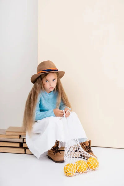 ファッショナブルなブロンド女の子で茶色の帽子とブーツ 白いスカートと青いセーター座っていますヴィンテージ本にグレープフルーツで文字列バッグ — ストック写真
