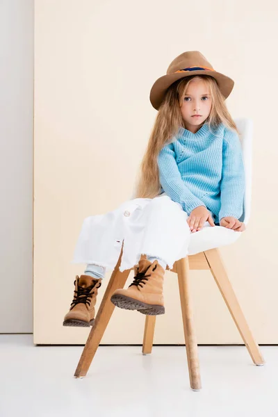 ファッショナブルなブロンドの女の子で茶色の帽子とブーツ 白いスカートと青のセーターはベージュの壁の近くの椅子に座って — ストック写真