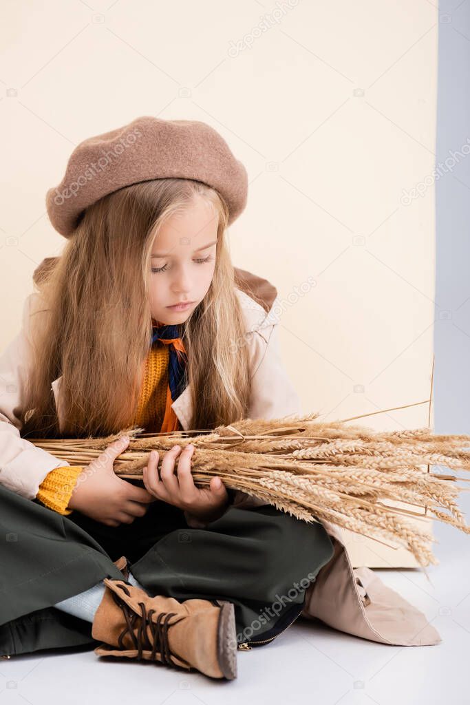 fashionable blonde girl in autumn outfit sitting on floor with wheat spikes on beige and white background